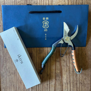 Ikiru secateurs with box and Tobisho gift bag – lifestyle photo showing secateurs with green and tan leather grip, Ikiru box, and Tobisho gift bag on a wooden surface.