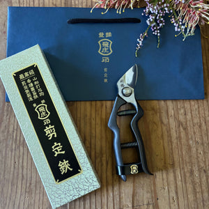 Tobisho Hirei Japanese pruning shears with gift box and branded blue bag, displayed on wooden surface with flowers