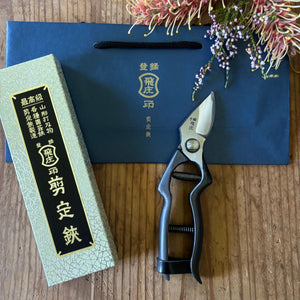 Tobisho Japanese pruning shears with black handles, gift box and branded blue bag on wooden surface with flowers