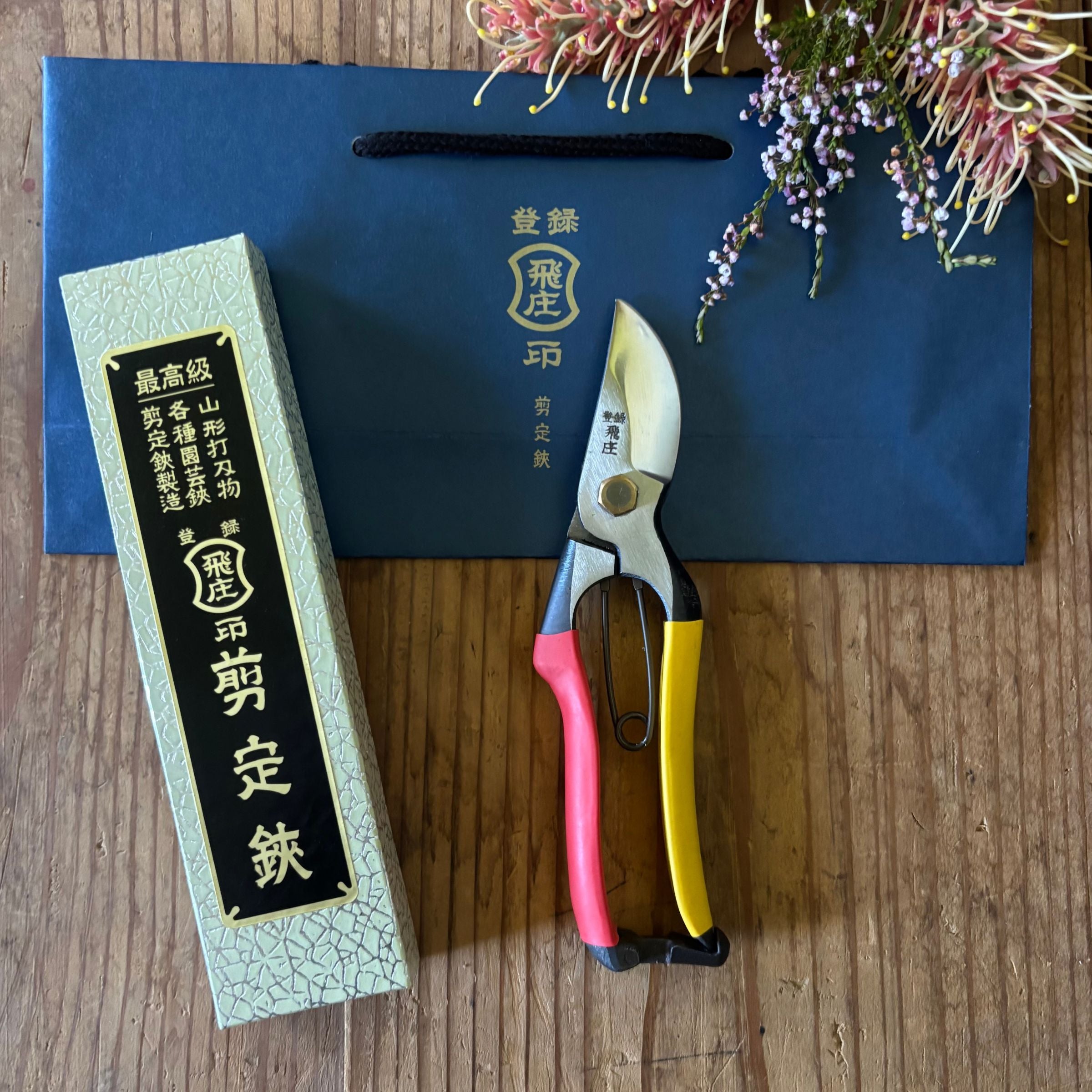Tobisho SR-1 pruning shears with red and yellow handles, gift box and branded blue bag on wooden surface with flowers