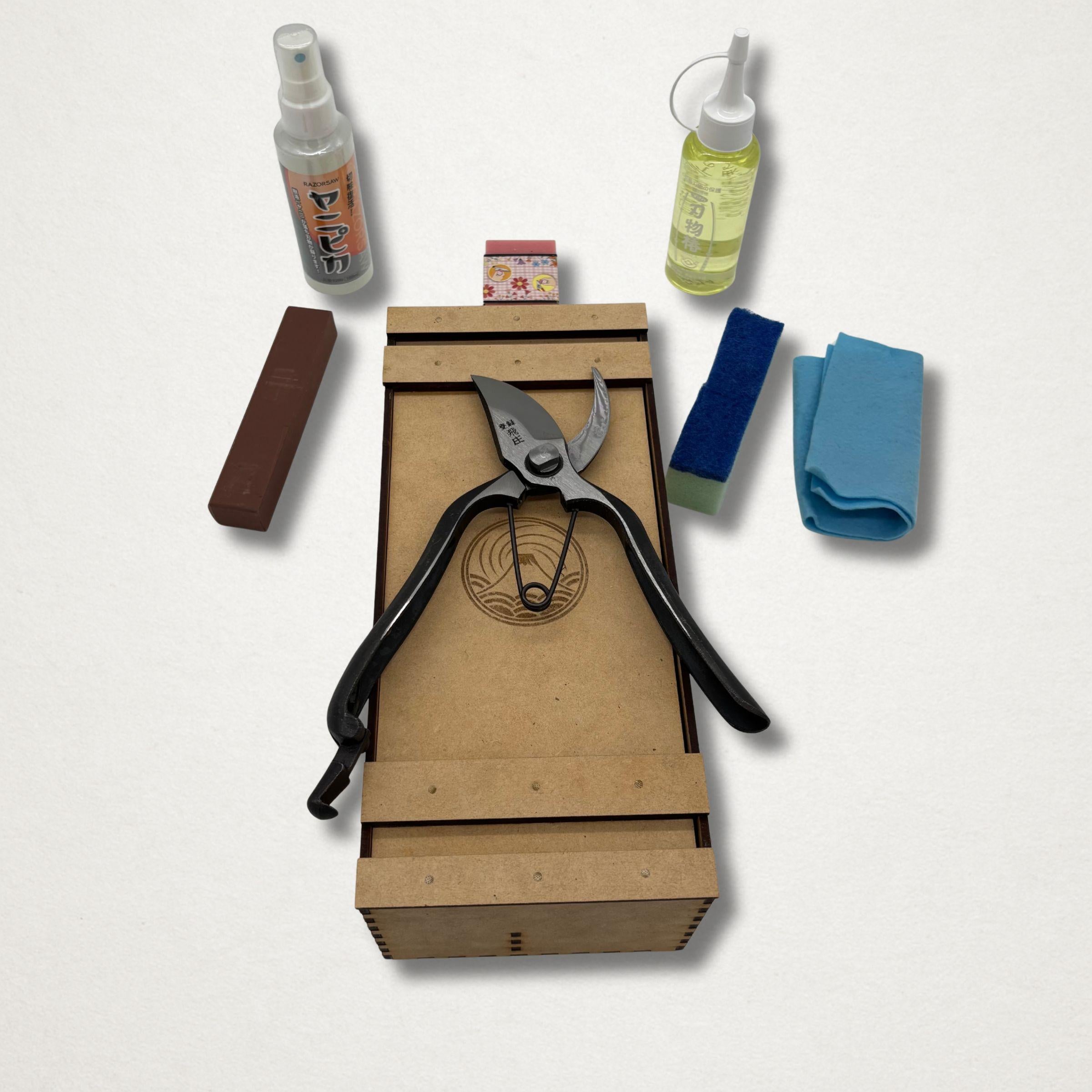 Tobisho Type B pruning shears stored on top of a handmade wooden toolbox, with blade cleaner, camellia oil, cloth, rust eraser and sharpening stone laid out beside it.