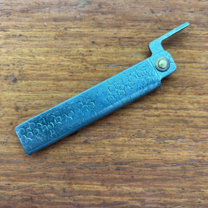 Japanese higo knife closed, showing hammered metal handle and thumb lever on a wooden surface