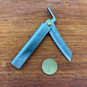 Japanese higo knife partly opened beside an Australian one dollar coin, showing hammered metal handle and compact blade on a wooden surface